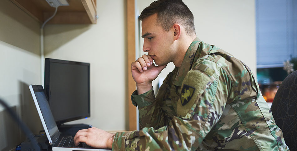 A service member in uniform sits at a desk, using a laptop.