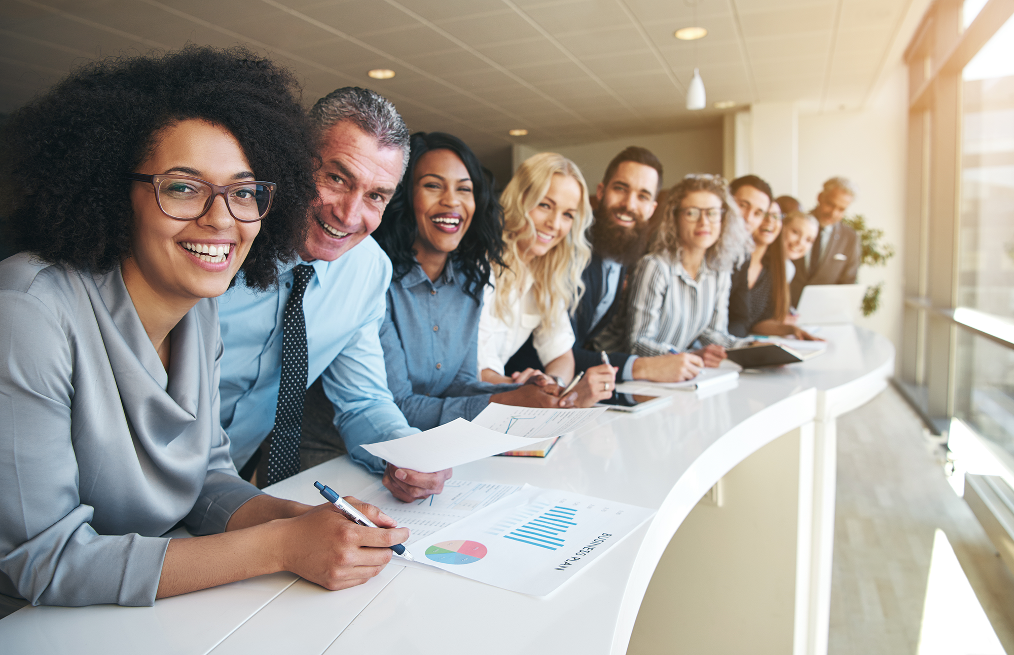 Row of people in business attire slightly bending over long conference table looking into camera.