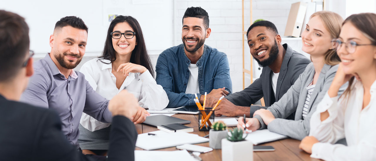 7 people sitting around a table having a meeting. Papers, phones, tablets, writing utensils are scattered around the table.