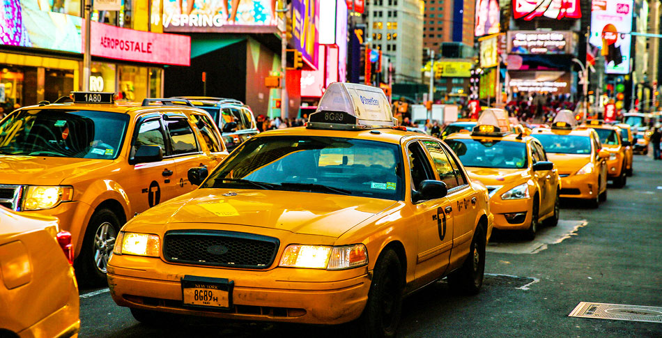 A street scene in New York city featuring taxi cabs and brightly lit storefronts and electronic billboards.