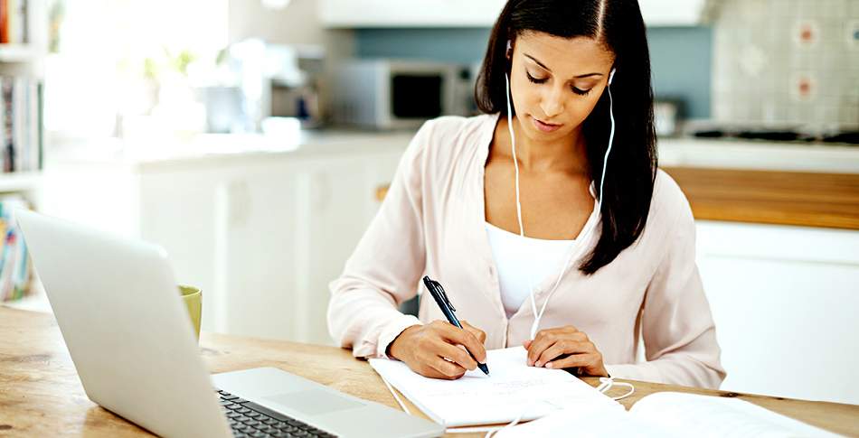 Woman with earbuds in and a laptop in front of her writing down notes on a notebook also in front of her.