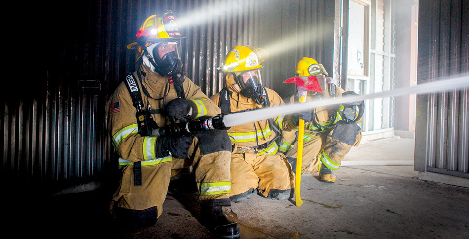 Three Firefighters in full gear inside of a building putting out a fire. Firefighter closest to the camera is holding a hose spraying water off screen.