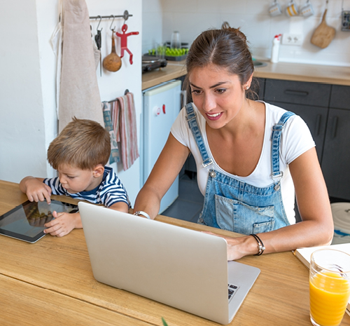 Woman sitting at a table looking at a laptop with a child sitting next to her looking at a tablet.