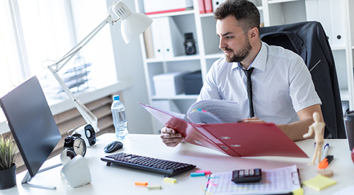 man sitting at a desk with a folder and calculator looking at his monitor