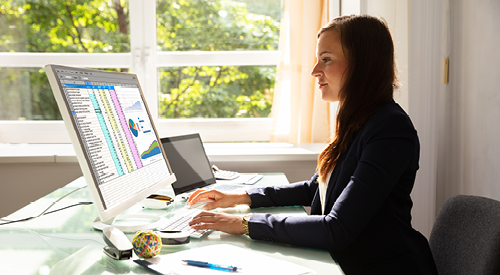 woman sitting at a desk with a spreadsheet and charts up on her monitor screen