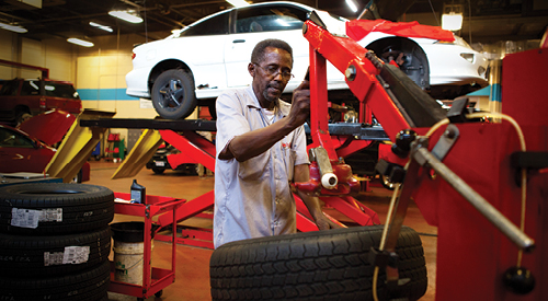 man working on putting a new rim inside of a tire with a car up on a jack behind him