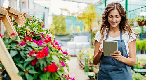 Woman holding a tablet inside of a greenhouse surrounded by potted plants and flowers