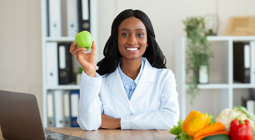 Woman holding an apple sitting at a desk. peppers and other vegetables are laying on the desk along with an open laptop