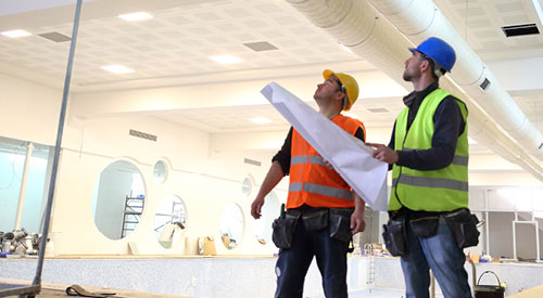 2 people looking up at ductwork wearing safety equipment holding building plans