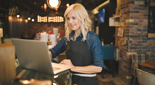 woman standing at a tablet looking at a laptop. restaurant décor in the background