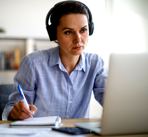 Woman sitting at a laptop using a pen to write on a notepad. She's wearing headphones.