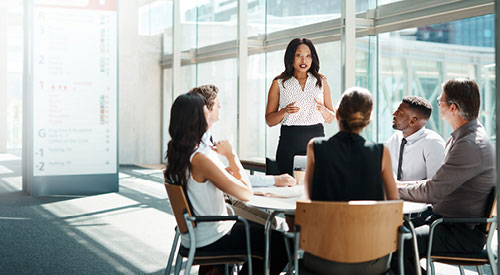 woman standing next to a conference table presenting while the other people sitting at the table look at her