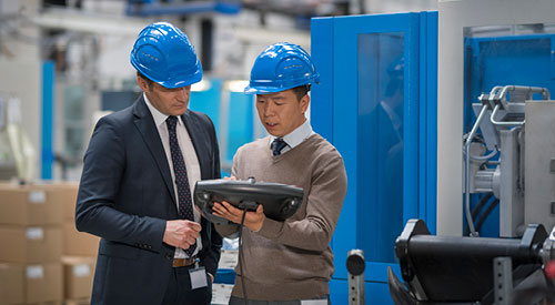 2 men wearing blue hard hats looking at a screen inside of a warehouse