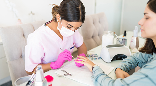 Woman with a mask and gloves applying nail polish to another woman sitting across from her.