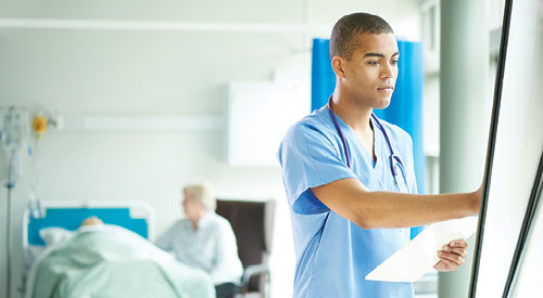 man in scrubs standing in front of a wall of charts. elderly woman seen in the background sitting next to a bed with an elderly man laying in it