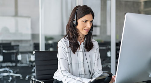 woman sitting at a desk in front of a computer wearing a headset attached to a phone