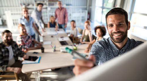 Man standing in front of a whiteboard while a room full of people sitting around a conference table behind him look at what he's drawing