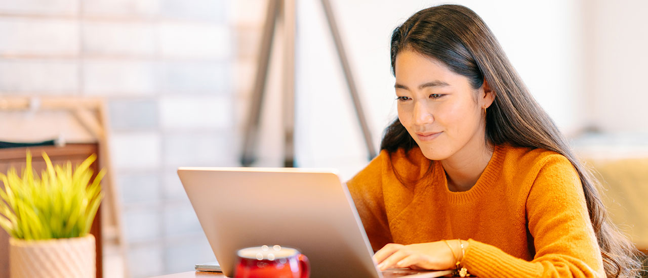 woman in orange sweatshirt sitting at a table working on a laptop