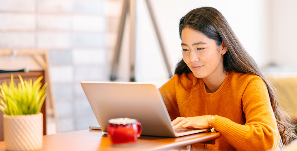 woman in orange sweatshirt sitting at a table working on a laptop