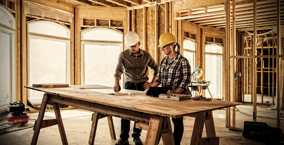 man and women wearing hard hats inside of a house being constructed looking at building plans laying on a piece of plywood.