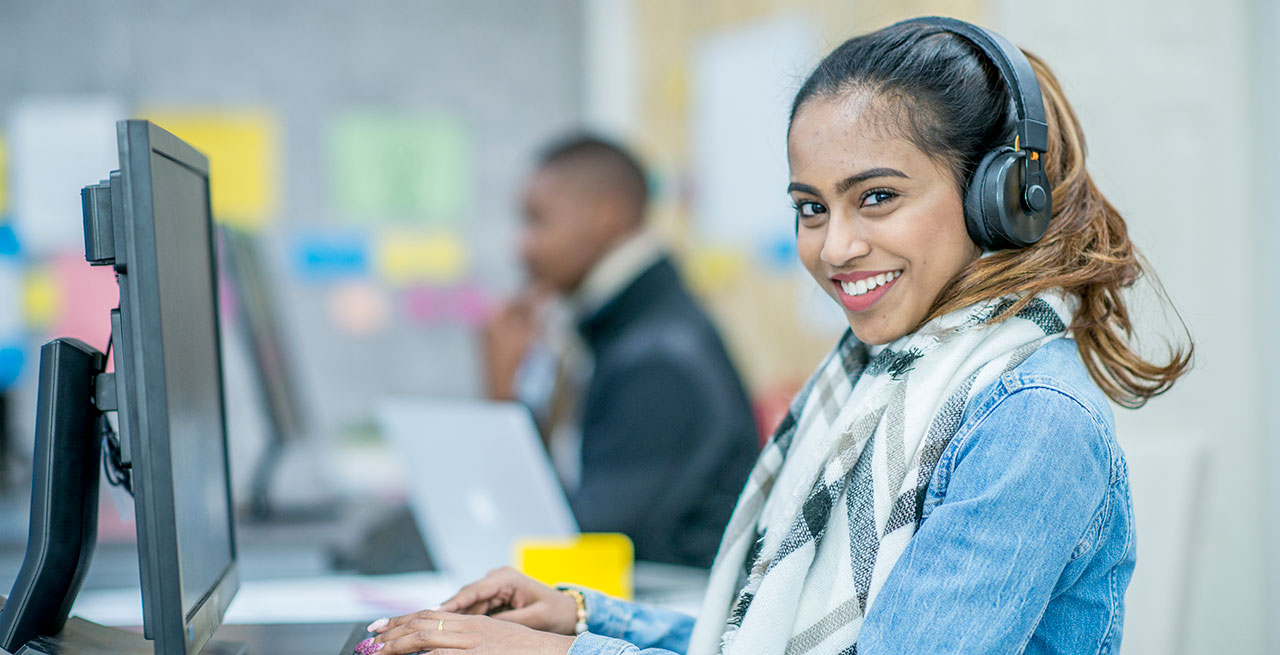 Woman sitting at a computer with a headset on looking at the camera smiling