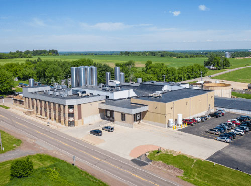 Nasonville Dairy's new building, a large, modern, multi-story structure with multiple stainless steel silos, cars in the parking lot and green fields in the distance.