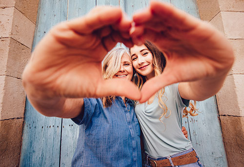 An older and younger woman stand arm-in-arm, forming a heart shape with their hands.