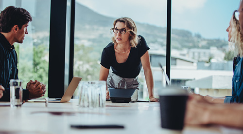 person standing at head of a conference table speaking to the other people sitting down at the table