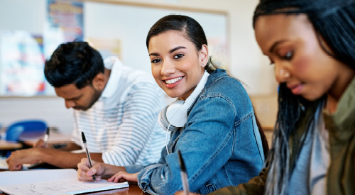 student looking at the camera holding a pen writing in a notebook a student is sat on each side of her