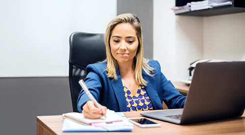 person sitting at desk writing on notepad with a laptop and phone next to them