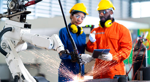 2 people standing back in hard hats and safety glasses observing a machine welding