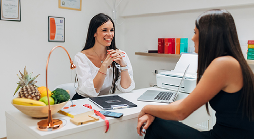 2 people sitting at a desk with a bowl of fruit on it talking