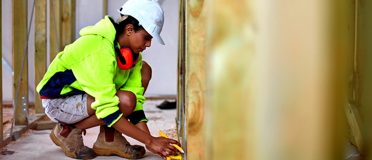 person squatting down in a building that is being constructed with a white construction hat and bright yellow sweatshirt measuring something on the floor 