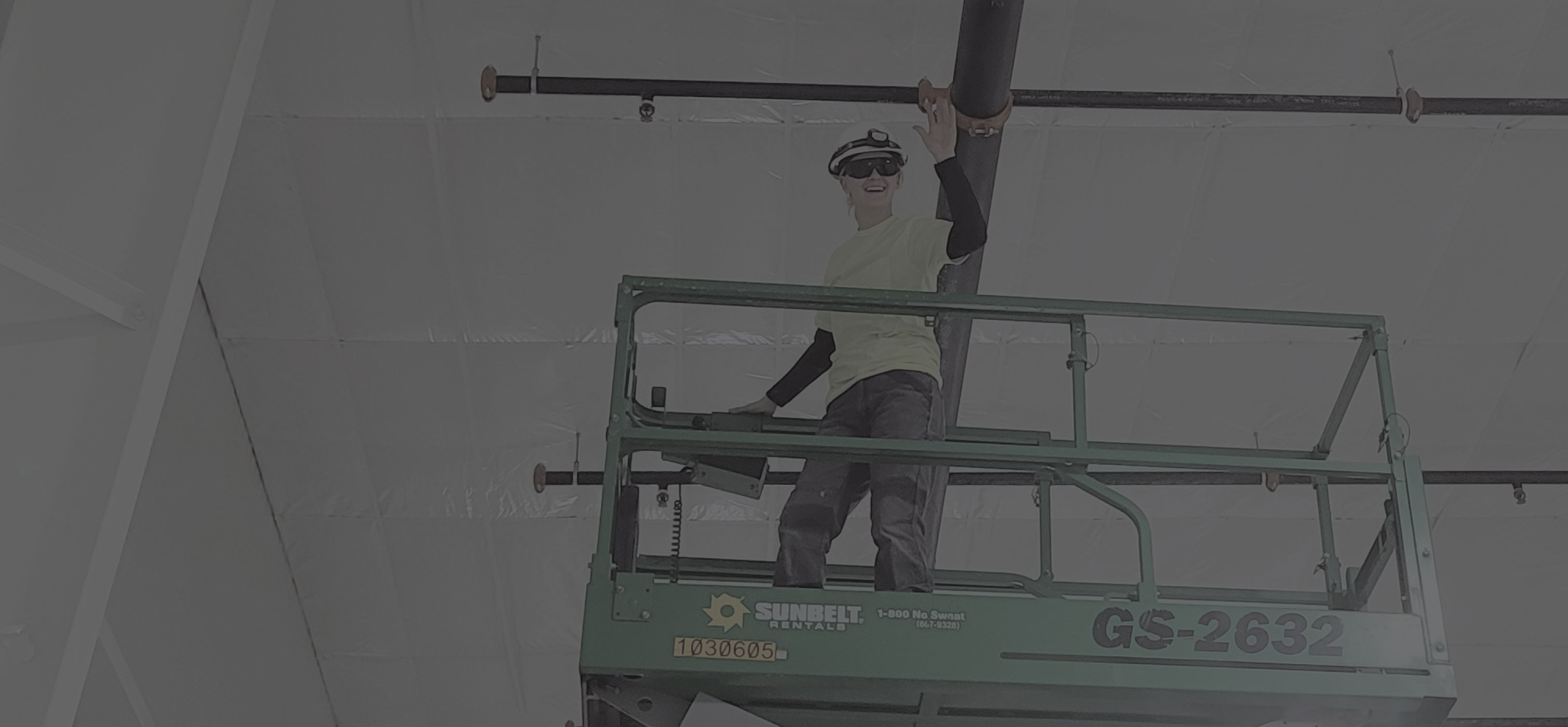 Woman standing on a lift, wearing a hard hat and safety glasses, waving.
