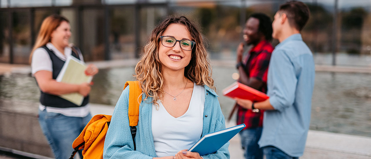 Person smiling at the camera wearing a backpack holding a book. Several people in the background having a conversation holding books.