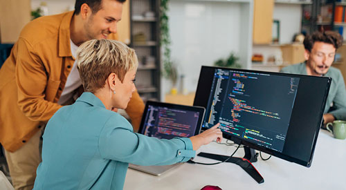 Person standing next to another person who is pointing to code on a monitor screen next to them