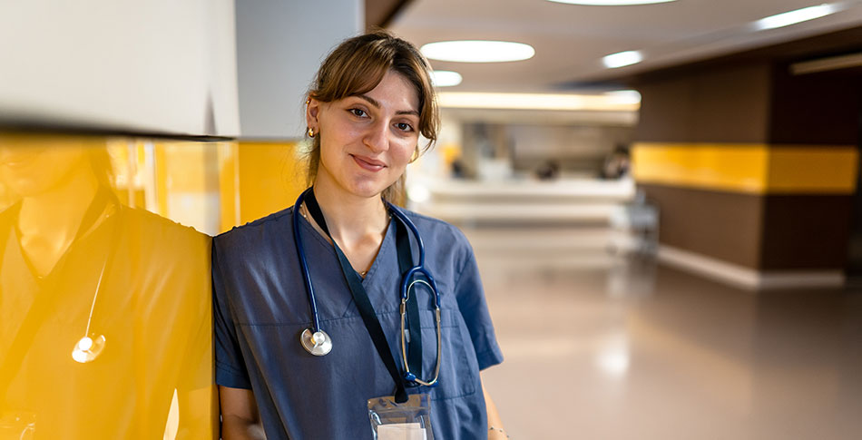 Nursing Assistant in blue scrubs leaning up against a reflective yellow wall.