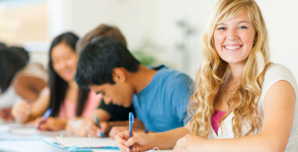 Person smiling at the camera while her peers write in notebooks next to her.