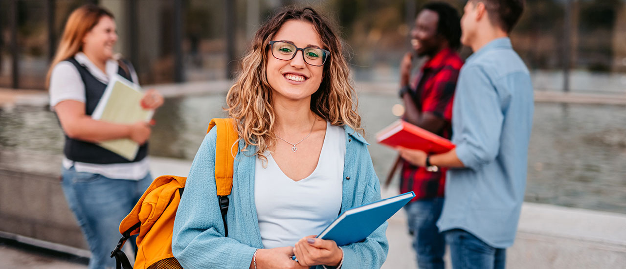 Student with a backpack on, holding a notebook smiling at the camera.