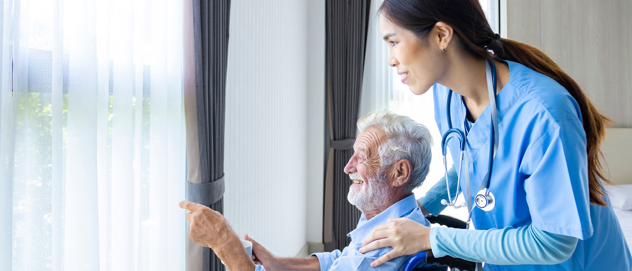 Nursing assistant stands behind man in a wheelchair while they look out the window.