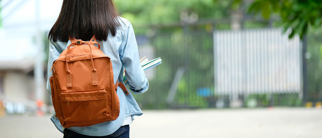 Woman wearing an orange backpack carrying books walking away from the camera.