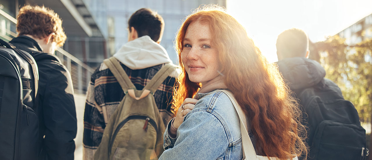 Group of 4 students with backpacks on walking toward a building. One student is turned around looking at the camera, sun shining in the background.