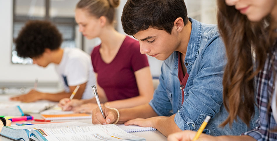 Students taking notes in notebooks while reading from a textbook.