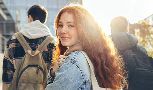 Group of 3 students with backpacks on walking toward a building. One student is turned around looking at the camera, sun shining in the background.