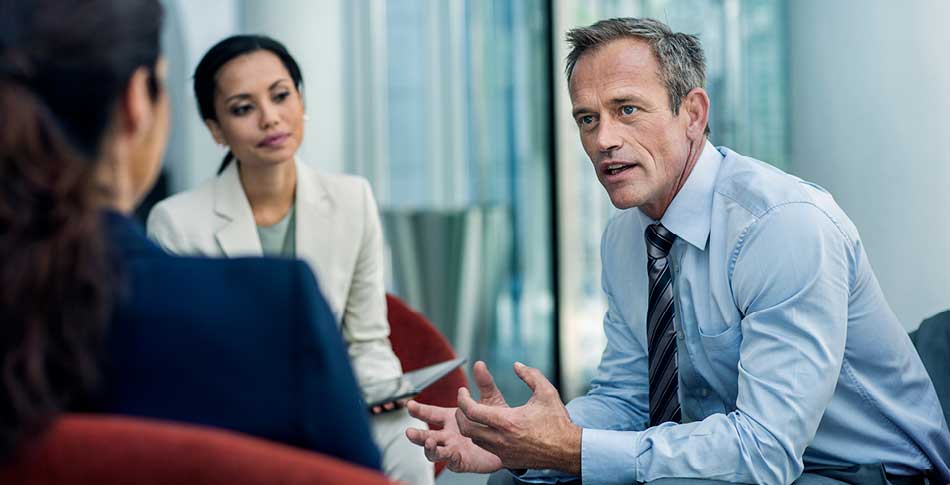 3 people sitting in chairs. in business attire. 2 of the people are looking at the 3rd person who is speaking.