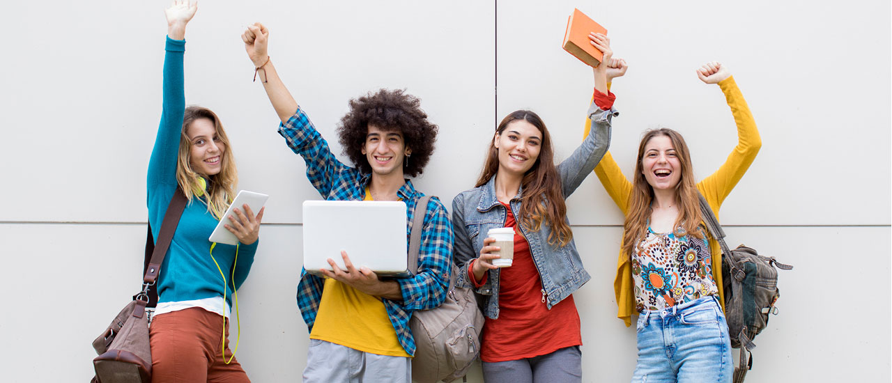 A group of four students standing together with backpacks, smiling and raising their arms in celebration.