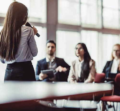 Person with a microphone standing in front of a group of people presenting.