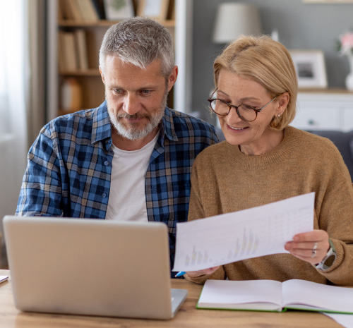 2 people sitting in front of a laptop. One is looking at the laptop the other is holding up a piece of paper they are looking at.