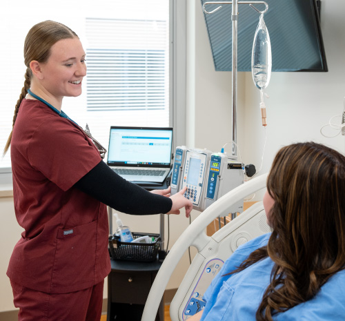 Person in red scrubs working with an IV bag next to a person sitting in a hospital bed