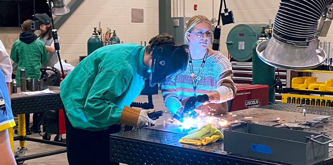A welding student in protective gear practices welding at a metal worktable while an instructor stands beside them, guiding the process in a busy welding lab.